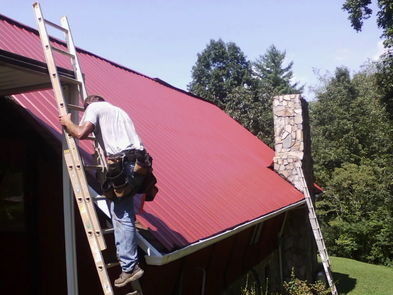 Roofer on a ladder beside a red metal roof and stone chimney — Soffit & Fascia Repair by Roofing Brothers in Chesnee